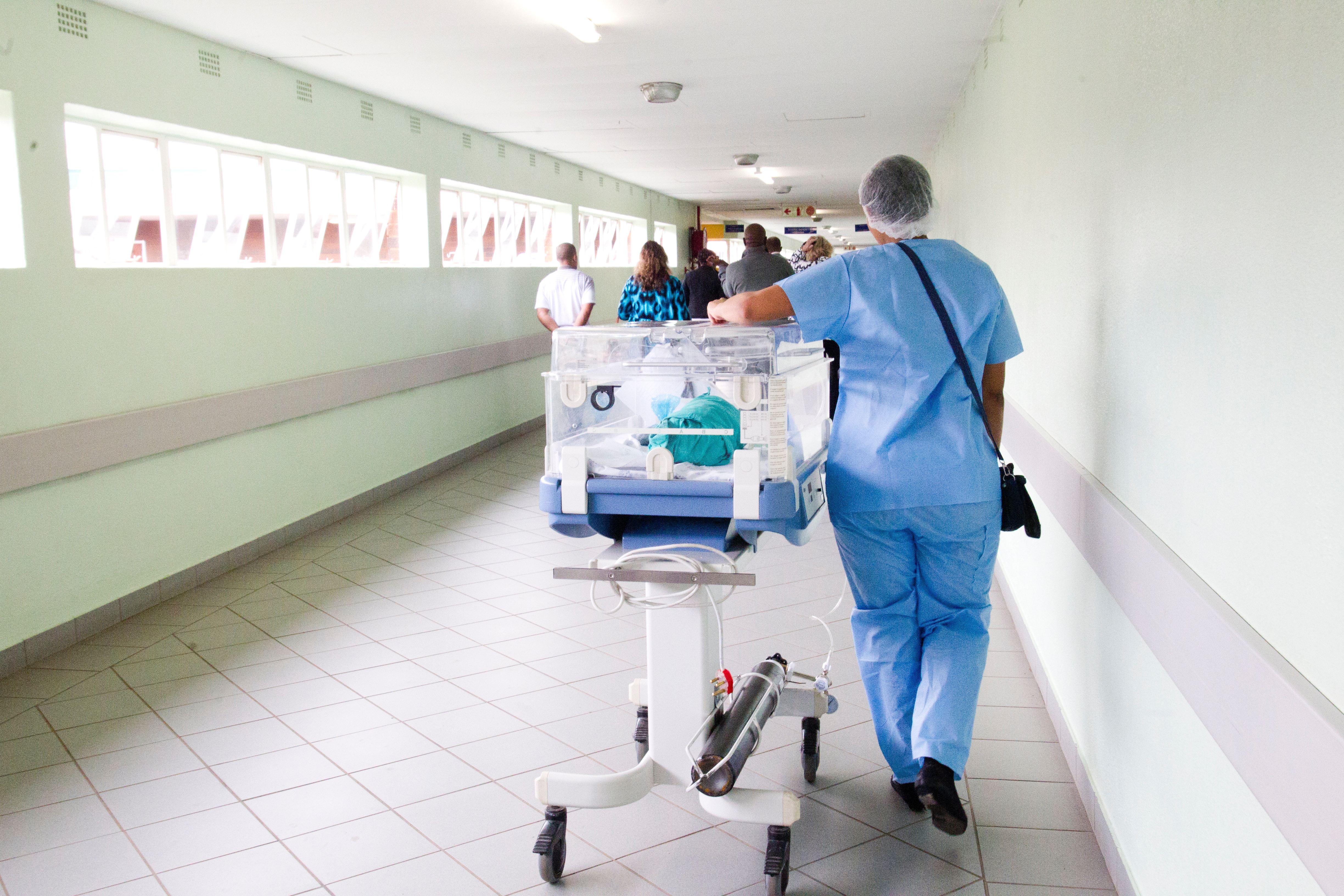 A nurse walking through a hospital hallway pushing along a container with a baby in it